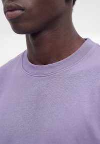 Close-up of a person's lower face and neck wearing a light purple crew neck shirt against a white background.