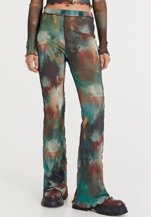 Close-up van een persoon die kleurrijke, met abstracte print versierde uitlopende broeken en bruine plateauzolen draagt, met zichtbare visnetmouwen zonder vingers.