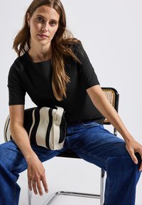 Haut noir à manches courtes avec un col carré, associé à un jean large bleu. Le modèle tient un sac rayé noir et écru.