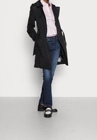 Persona vestita con un trench coat nero, camicia bianca con bottoni, jeans blu e mocassini bianco e nero, in piedi davanti a uno sfondo chiaro e semplice.