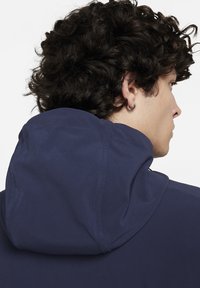 Hooded sweatshirt in marineblauw stof. Heeft een gladde textuur, zichtbare stiksels en een capuchon met trekkoord. Achteraanzicht toont haar en een oorbel.
