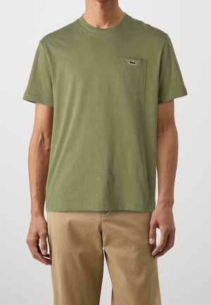 Homme portant un T-shirt olive à manches courtes avec une poche poitrine et un petit logo crocodile, associé à un pantalon beige, sur un fond uni.