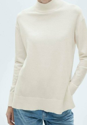 Vrouw draagt een crèmekleurige coltrui met lange mouwen en een blauwe spijkerbroek, met één hand in de zak.