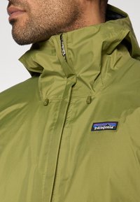 Πράσινο αδιάβροχο μπουφάν με ψηλό γιακά, φερμουάρ και ρυθμιζόμενα κουμπώματα. Διαθέτει μαύρο λογότυπο Patagonia στο αριστερό στήθος.