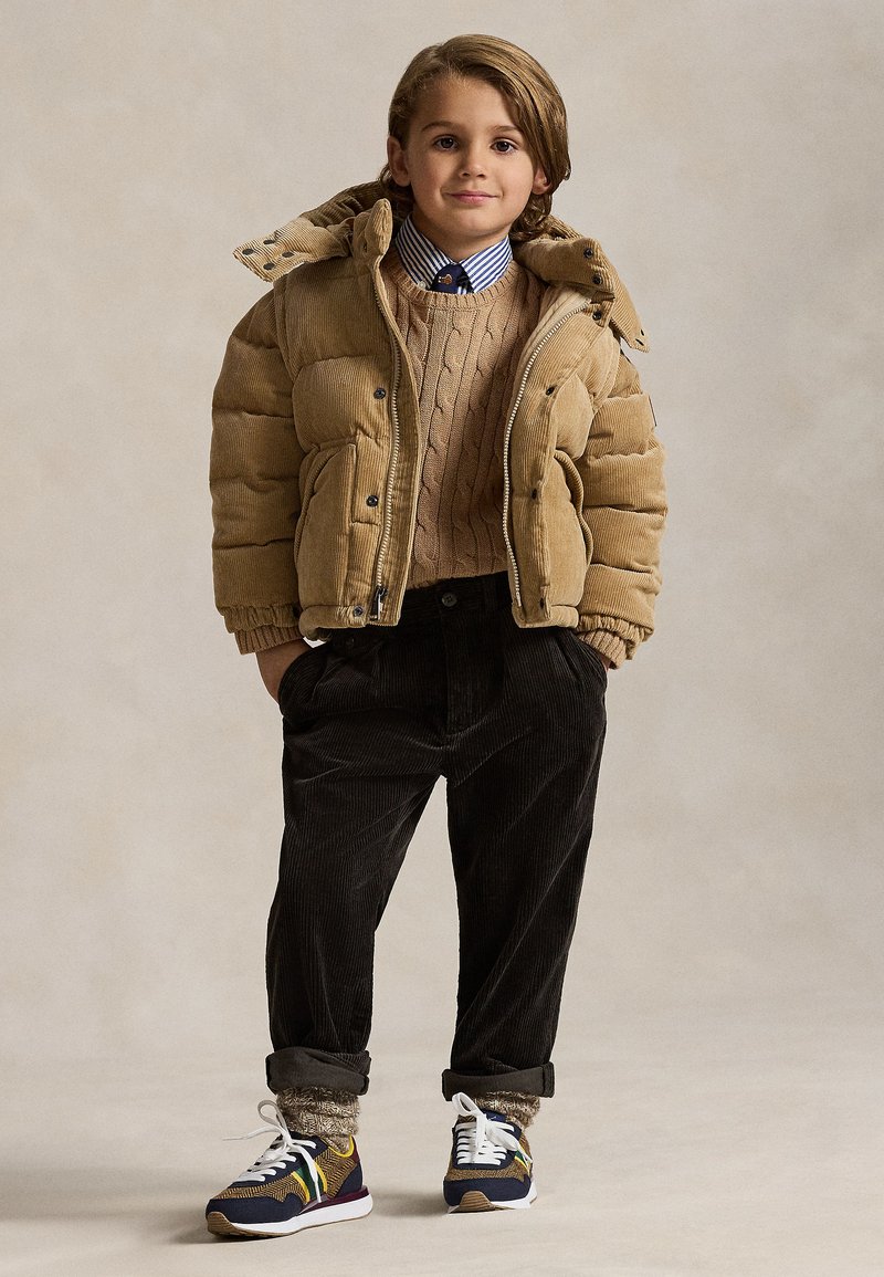 Ragazzo giovane in piedi con le mani nelle tasche, indossa una giacca imbottita beige, un maglione color cammello, una camicia a righe con cravatta, pantaloni scuri, calze a fantasia e scarpe da ginnastica colorate.