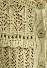 Tissu en maille beige avec des motifs géométriques et en dentelle, présentant un bord festonné et trois boutons en plastique le long d'un panneau côtelé.