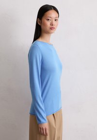 Helles blaues Langarmshirt mit rundem Halsausschnitt, glatter Textur und abgerundetem Saum. Kombiniert mit beigen Hosen für den Kontrast.