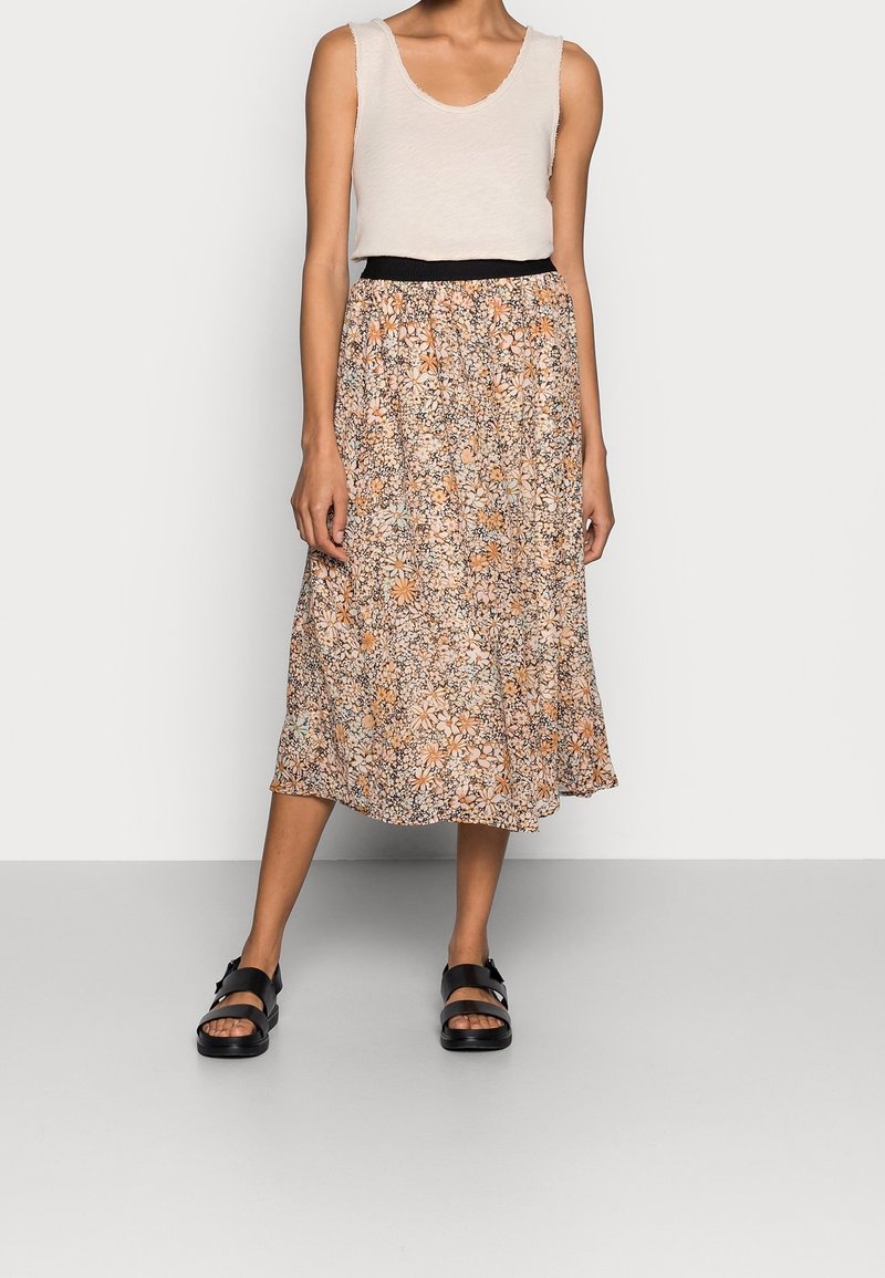 Femme portant un haut beige sans manches, une jupe fleurie mi-longue avec une ceinture noire, et des sandales noires, debout devant un fond blanc.