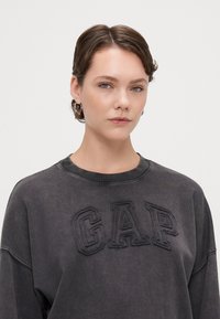 Sudadera gris con un logo "GAP" bordado, cuello redondo y un ajuste ligeramente oversize. Tejido suave con una textura lisa.