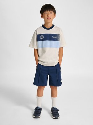 Dreng stående med hænderne i lommerne, iført hvid og blå sportstrøje, marineblå shorts, hvide sokker og marineblå sneakers mod en ensfarvet baggrund.