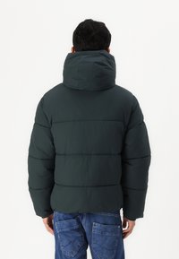 Tamnozeleni puffer jakna s visokim ovratnikom, koja ima horizontalne kvadrate i opušteni kroj. Uparen s plavim trapericama.