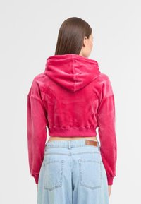 Gekürzter rosa Hoodie mit einer weichen Textur und Kapuze, ausgestattet mit gerippten Bündchen und Saum. Kombiniert mit hellblauen, hoch geschnittenen Jeans.