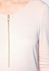 Jasnoróżowa bluzka z okrągłym dekoltem i widocznym zamkiem błyskawicznym z przodu. Materiał wydaje się gładki z subtelnym połyskiem.