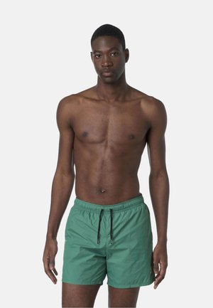 Giovane uomo con pelle scura, capelli corti, che indossa pantaloncini da bagno verdi con coulisse nere, in piedi davanti a uno sfondo chiaro e uniforme.