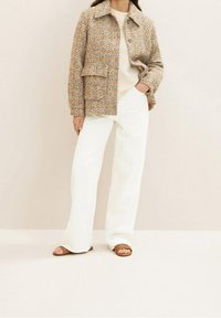 Vrouw in een bruine en grijze gestructureerde jas over een lichtgestreept topje, witte wijde broek en bruine open-toe sandalen.