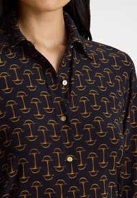 Tuvu-plāns melnas krāsas krekls ar brūnu atkārtotu rakstu un zelta pogām, ko valkā persona ar gariem tumšiem matiem.