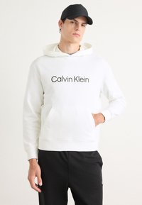 Hanorac alb cu buzunar frontal și imprimat "Calvin Klein" în gri. Purtați cu pantaloni negri. Dispune de o glugă cu șnur și manșete cu textură ribbed.