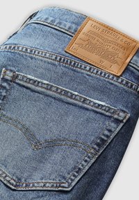Jean en denim bleu avec une poche arrière cousue et un patch en cuir marron de la marque Levi Strauss & Co. au-dessus de la poche.