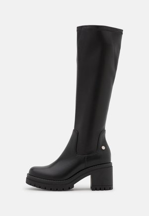 Botte haute en cuir noir avec talon bloc épais et semelle crantée, tige sans couture et petit logo métallique rond sur le côté près de la cheville.