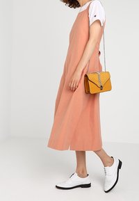 Abito in lino color pesco con una vestibilità morbida, abbinato a una maglietta bianca, una borsa gialla con catena in argento e scarpe brogue bianche.