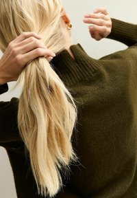 Olivgrüner Pullover mit geripptem Kragen, getragen von einer Person, die lange, lockere, blonde Haare zurückbindet. Der Fokus liegt auf der Textur und Farbe.