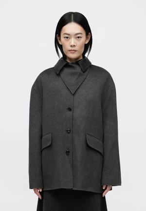 Manteau en laine grise avec un col en V, des accents en cuir noir et de grandes poches avant. Présente une silhouette décontractée et une fermeture à boutons.