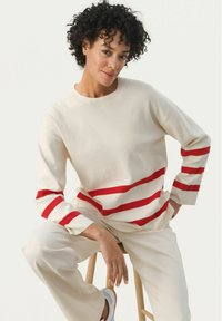 Kermanharma sweatshirt, jossa on punaiset vaakasuorat raidat hihoissa ja vartalossa, valmistettu pehmeästä kankaasta, yhdistettynä yhteensopiviin vaaleisiin housuihin.