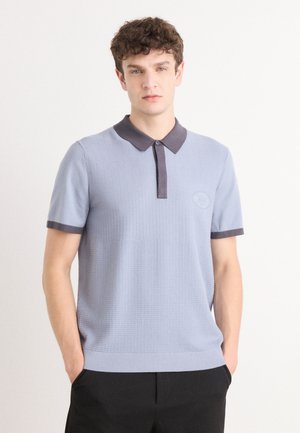 Lys blå strikket polo skjorte med mørk krage og mansjetter, med en subtil tekstur og en rund logo på brystet.