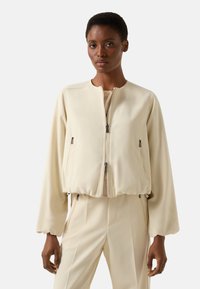 Kremowa kurtka bomberka z dopasowanym pasem, wyposażona w przedni zamek błyskawiczny i dwie boczne kieszenie na zamek. Gładka faktura i długie rękawy.