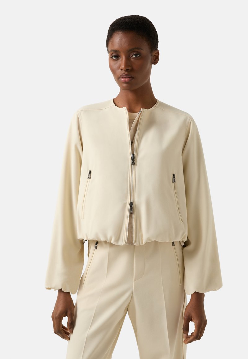 Kremowa kurtka bomberka z dopasowanym pasem, wyposażona w przedni zamek błyskawiczny i dwie boczne kieszenie na zamek. Gładka faktura i długie rękawy.