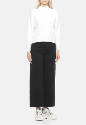 Maglione a dolcevita bianco, design corto, indossato con jeans neri a gamba larga e sneaker bianche con suola platform, su sfondo grigio chiaro.