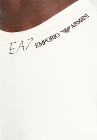 White cotton sweatshirt with a ribbed neckline. Features brown embroidered "EA7" and "Emporio Armani" logos on the collar.