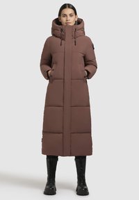 Hosszú barna párnázott kabát magas gallérral, levehető kapucnival, két elülső zsebbel és egyenes szabással, fekete csizmákkal párosítva.