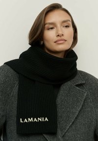 Femeie îmbrăcată cu o eșarfă neagră cu textul "LAMANIA" în jurul gâtului și cu un palton gri cu model herringbone, pe un fundal simplu.