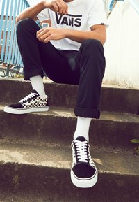 Baskets Vans à damier noir et blanc avec lacets blancs, portées avec des chaussettes blanches et un pantalon noir retroussé, posées sur des marches en béton.