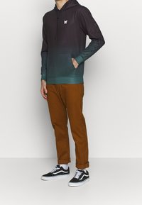 Uomo che indossa un hoodie ombre nero-verde con un logo a farfalla, pantaloni marroni risvoltati, calze bianche e sneaker bianco e nere.