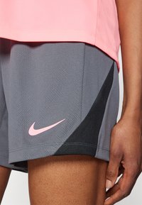 Nike atletske kratke hlače u sivoj boji s teksturiranom površinom. Imaju crne naglaske i ružičasti Nike logo na donjoj lijevoj strani.