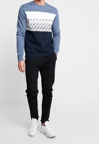 Mikina v modré, bílé a námořnické modři s vodorovnými barevnými bloky a logy. Kombinováno s černými tepláky a bílými teniskami. Měkká látka, volný střih.