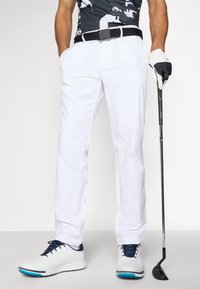 Hvite golfbukser med en glatt tekstur, svart belte, og en poloskjorte med svart og hvit mønster. Hvite golfskobr med marineblå detaljer. Golfkølle i hånden.