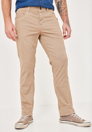 Beige cotton pants with a textured finish, featuring front pockets, a button closure, and paired with grey canvas sneakers.