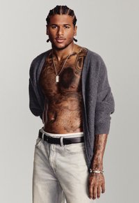 Gray cardigan, muscular torso displaying intricate tattoos, light denim jeans, black belt, and a silver bracelet. Simple background.