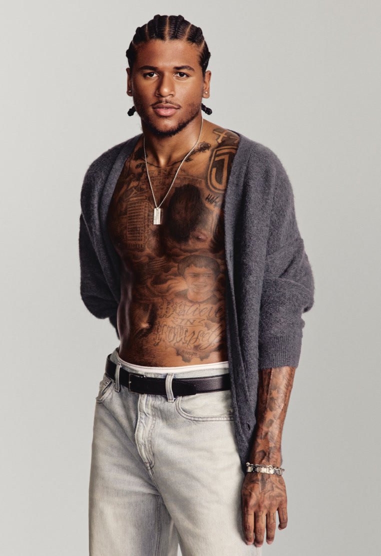 Gray cardigan, muscular torso displaying intricate tattoos, light denim jeans, black belt, and a silver bracelet. Simple background.