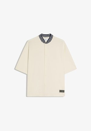 Väriltään luonnonvalkoinen lyhythihainen paita, jossa painonapillinen etumppu, mustavalkoraidallinen kaulus ja pieni musta suorakaiteen muotoinen logopläjäys helmassa.