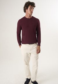 Burgundy long-sleeve crew neck sweater with ribbed cuffs, paired with light-colored trousers and dark sneakers. Simple, textured fabric.