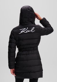 Abrigo acolchado negro con capucha, cintura ajustada, mangas largas y texto blanco bordado "Karl" en la parte superior de la espalda. Tejido texturizado y brillante.