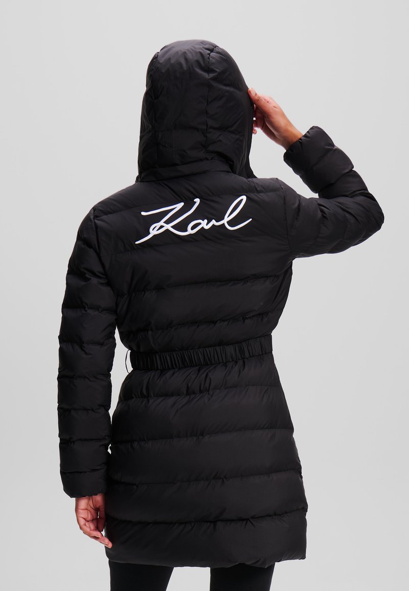 Abrigo acolchado negro con capucha, cintura ajustada, mangas largas y texto blanco bordado "Karl" en la parte superior de la espalda. Tejido texturizado y brillante.