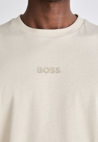 Beige T-skjorte med rund hals laget av mykt stoff, med preget "BOSS"-logo i matchende farge på brystet. Glatt tekstur, avslappet passform.