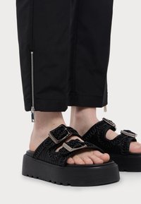 Schwarze Plateausandalen mit strukturierten Riemen und metallischen Schnallen, kombiniert mit schwarzen Hosen, die am Saum einen silbernen Reißverschluss haben.