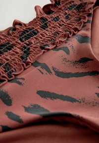 Rostfärgad tyg med svarta abstrakta penseldragmönster, med en rynkad detalj upptill längs kanten. Slät textur.