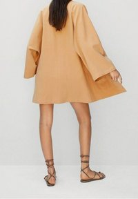 Beige oversized kaapu with broad sleeves, crafted from textured fabric, worn over brown strappy sandals. The silhouette is loose and flowing.